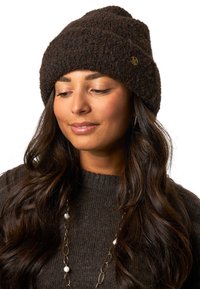 Brązowa dziana czapka z grubym, teksturowanym splotem i lekko podwiniętym brzegiem. Posiada małe złote logo z boku.