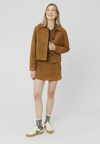 Veste courte en daim marron avec poche avant et boutons, mini-jupe en daim assortie, baskets crème et chaussettes imprimé léopard.