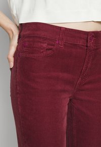 Bordeaux corduroy broeken met verticale ribbels, voorzien van twee achterzakken en paarse knopen op de tailleband. Zachte textuur en aansluitend ontwerp.