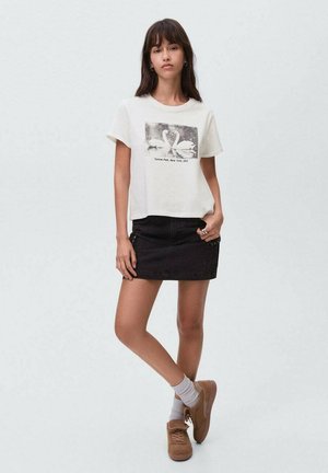 T-shirt blanc court avec un motif de cygne noir, associé à une jupe en denim noir. Des baskets marron et des chaussettes grises complètent la tenue.