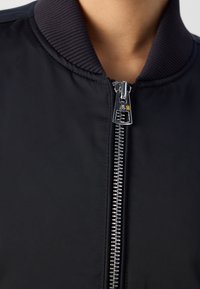 Černá bomber bunda s žebrovaným límcem, která má stříbrný zip a hladkou, mírně lesklou tkaninu. Detailní záběr na texturu látky a detaily zipu.