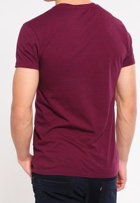 Homme portant un t-shirt bordeaux ajusté à manches courtes et un jean bleu foncé, vu de dos.