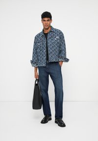 Denim jakna z geometrijskim vzorcem, modri kavbojki, črna majica, drži črno torbo, črne čevlje, stoji ob nevtralni ozadju.