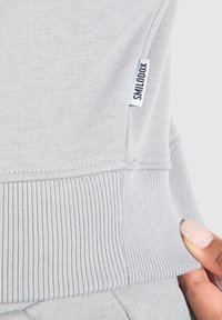 Lys grå sweatshirt med ribbet kant og manchetter, glat tekstur og et lille mærke på siden til identifikation.
