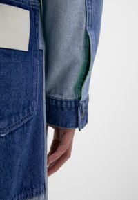 Jeansjacka med färgblockdesign i mörkblå och ljusblå sektioner, knäppt ärmslut och gröna detaljer på insidan av ärmen.