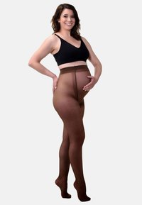 Collant da maternità marroni con una texture liscia e trasparente, progettati per offrire supporto. Indossati con un bralette nero, che garantisce una vestibilità aderente attorno alla pancia.