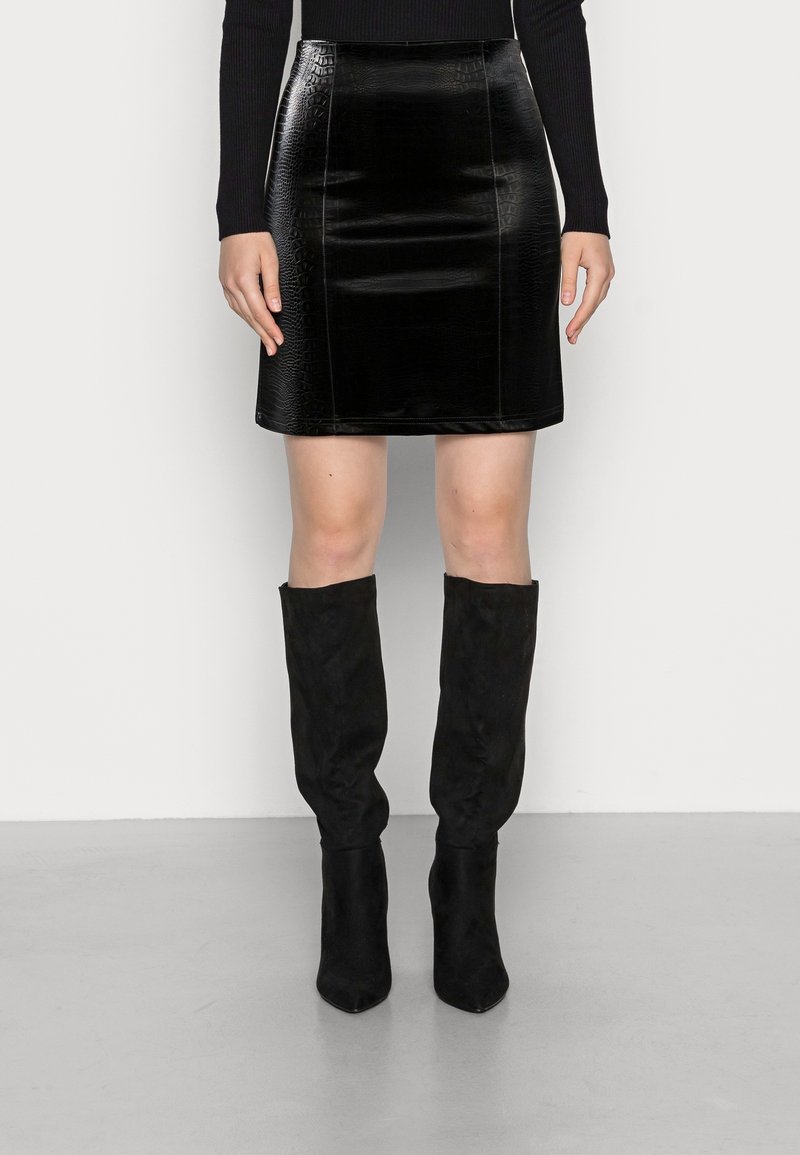 Femme portant une mini-jupe noire texturée, un haut noir à manches longues et des bottes noires en daim montant jusqu'aux genoux, debout sur un sol gris.