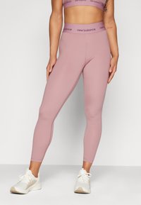 Ženska oblečena v visoko pasne mauve New Balance leggins hlače in ujemajoči športni modrček z belimi športnimi čevlji, stoji ob preprosti ozadju.