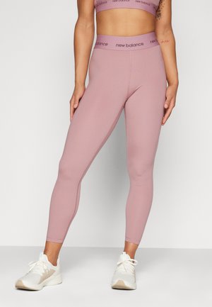 Donna che indossa leggings mauve a vita alta di New Balance e un reggiseno sportivo abbinato, con scarpe da ginnastica bianche, in piedi su uno sfondo semplice.