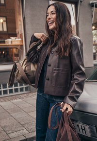 Brun läderjacka med en rak passform, knappstängning och två framfickor, kombinerad med mörka jeans och en beige axelväska.