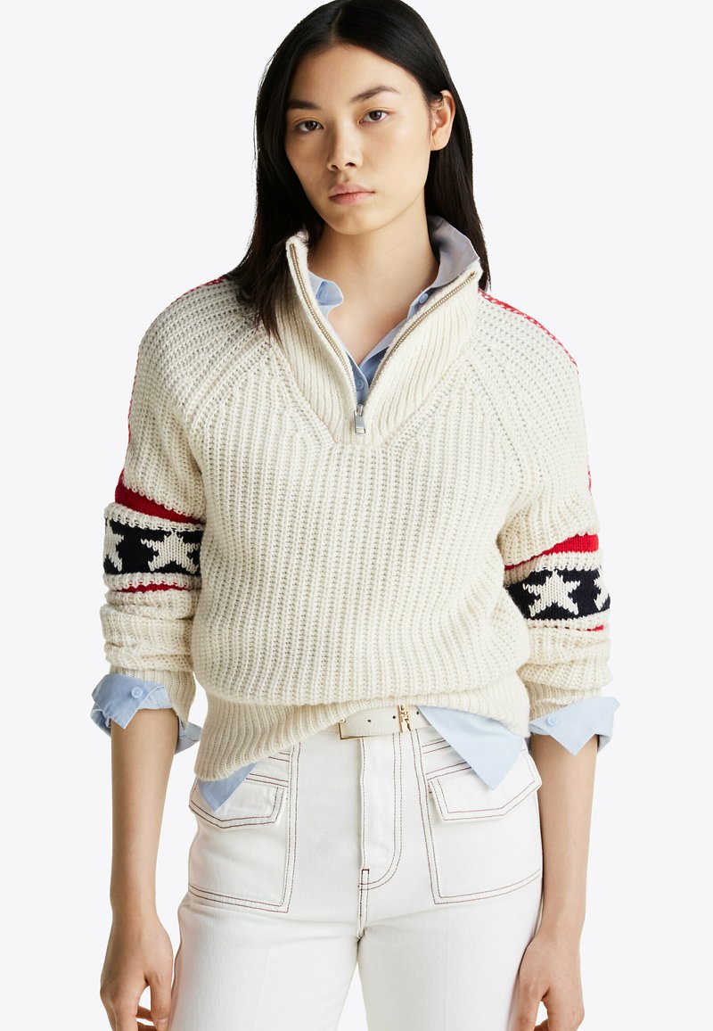 Kremowy sweter z kroszonką i zamkiem ćwierć, z czerwono-czarnymi wzorzystymi rękawami, noszony na jasnoniebieskiej koszuli z guzikami, w zestawie z białymi spodniami.