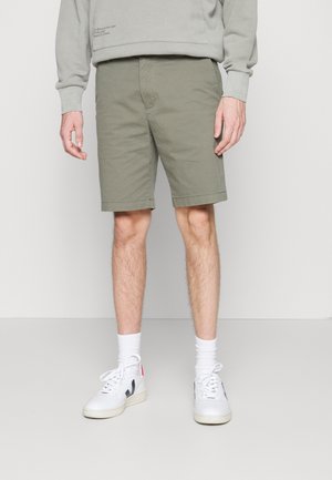 Pantalones cortos de algodón verdes con bolsillos delanteros, un ajuste recto y un dobladillo justo por encima de la rodilla, combinados con zapatillas blancas y calcetines hasta el tobillo.