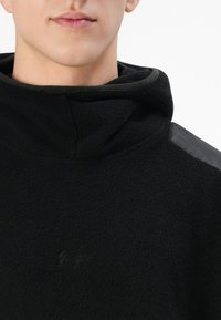 Primo piano di una persona che indossa un hoodie in pile nero con un piccolo logo ricamato e un pannello in nylon sulla spalla, su sfondo bianco.