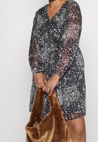Vestido negro con flores y mangas largas transparentes, cintura ajustada y escote en forma de V. Sosteniendo un bolso de piel de pescado marrón suave, que muestra un delicado patrón floral.