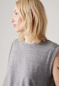 Blonde woman with shoulder-length hair wearing a sleeveless gray top, facing left, showing small tattoos on her upper arm.