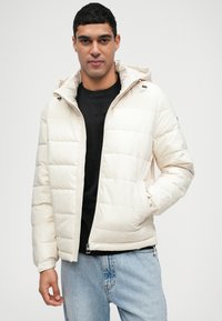 Κρέμ puffer τζάκετ με φερμουάρ και ελαστικούς μανσέτες, διαθέτοντας κουκούλα και ραμμένο σχέδιο. Φοριέται πάνω από ένα μαύρο πουκάμισο.