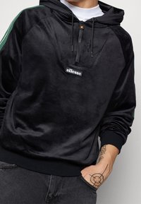 Felpa nera in velluto con mezza zip, strisce laterali verdi e logo ricamato sul davanti. Maniche e orlo a costine.