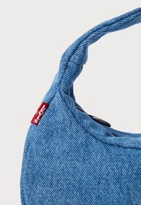 Denim handtas in lichtblauw, met een gestructureerde halve maanvorm, een stoffen handvat en een rood merketiket met een knoopdetail.