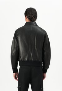 Hombre con chaqueta de cuero negra y pantalones negros, de pie de espaldas a la cámara contra un fondo blanco liso.