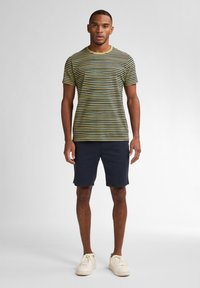 T-shirt rayé jaune et vert foncé, col rond, associé à un short bleu marine. Modèle portant des baskets couleur crème.