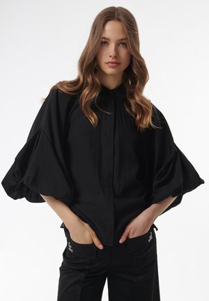 Camisa negra con cuello, que presenta mangas abullonadas y voluminosas, y una sutil textura vertical. Combinada con pantalones negros ajustados con botones en los bolsillos.