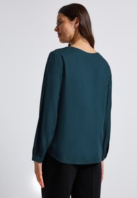 Blusa teal a maniche lunghe, con leggera lucentezza, vestibilità ampia, scollo rotondo e dettagli arricciati sui polsini; abbinata a pantaloni neri.