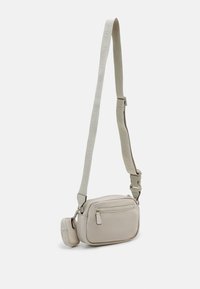 Beige crossbody taske lavet af glat materiale med en lynlåslomme foran og en justerbar rem med præget branding.
