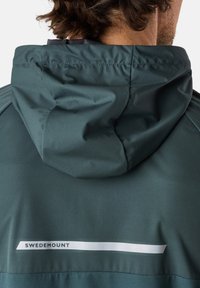 Giacca verde scuro con cappuccio, realizzata in materiale liscio. Una striscia di logo bianca con il testo "SWEDEMOUNT" è visibile sul retro.