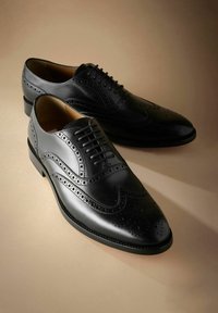 Een paar zwarte leren brogues met wingtip-detail, geplaatst op een beige oppervlak met zacht licht.