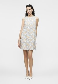Žena v šatách bez rukávov po kolená s jemným kvetinovým vzorom v svetlomodrej, béžovej a bielej farbe, v kombinácii s bielymi lodičkami so špicatou špičkou.