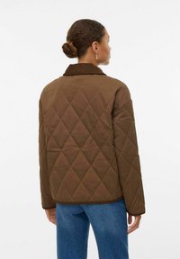 Veste matelassée marron avec un motif en losanges, col en velours côtelé et manches longues. Portée avec un jean en denim bleu, vue de dos.