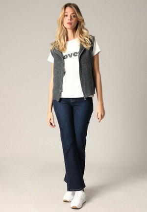 Grijze mouwloze vest over een wit t-shirt met zwarte 'love' tekst, gecombineerd met donkerblauwe uitlopende jeans en witte sneakers. Neutrale achtergrond.