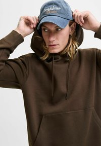Brown hooded sweatshirt with drawstrings and a front pocket, paired with a blue cap featuring white embroidered logo. Casual design.