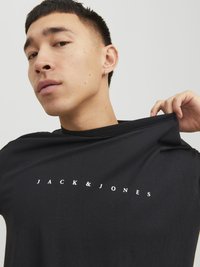 Schwarzes T-Shirt mit "JACK & JONES" in weißem Text. Der Stoff wirkt weich, hat eine lockere Passform und eine leicht strukturierte Oberfläche.