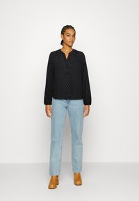 Blusa preta de mangas compridas com bolinhas brancas, frente com botões, decote em V e punhos elásticos, combinada com jeans azul claro e sapatos de salto bege.
