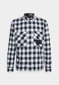Camicia in flanella a quadri bianchi e neri con colletto button-down, due tasche frontali e un accento nero solido sulla tasca destra.