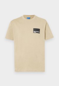 Vaahto beige puuvillapaita lyhyillä hihoilla, pyöreällä pääntiellä ja mustalla suorakulmaisella taskulla, jossa on valkoisella painettuna "KARL LAGERFELD JEANS".