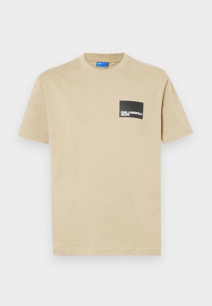 Beige pamut póló rövid ujjakkal, kerek nyakkal, és egy fekete téglalap alakú zsebbel, amelyen fehér betűkkel a "KARL LAGERFELD JEANS" felirat található.