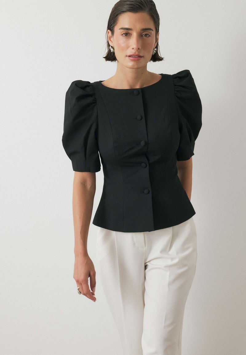 Top peplum nero con maniche a sbuffo e sei bottoni rotondi davanti, abbinato a pantaloni bianchi, su uno sfondo neutro.
