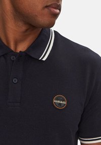 Polo shirt blu navy con strisce bianche sul colletto, caratterizzato da un logo circolare arancione e nero sulla zona sinistra del petto.