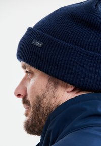 Navy sininen neulepipo, jossa on teksturoitu ribbikuvio ja pieni musta logotäydennys sivussa, käytetty tumman takin kanssa.