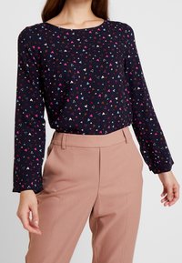 Blusa navy con maniche lunghe caratterizzata da un motivo di cuori multicolori. Abbinata a pantaloni su misura rosa chiaro. Tessuto con texture liscia.