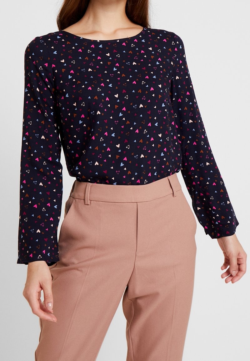 Blusa navy con maniche lunghe caratterizzata da un motivo di cuori multicolori. Abbinata a pantaloni su misura rosa chiaro. Tessuto con texture liscia.