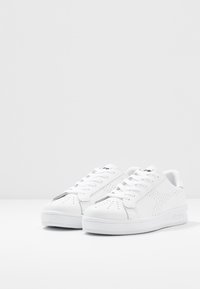 Sneakers atletiche bianche realizzate in materiale sintetico, con perforazioni sui lati, lacci piatti e suola in gomma. Design semplice senza motivi.