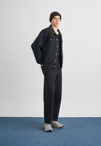 Casaco de ganga preto e jeans combinando, acompanhados por ténis cinzentos. Modelo usa um gorro castanho. Design simples, com detalhes minimalistas.