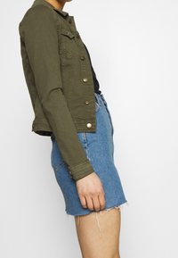 Olivgrüne Jeansjacke mit Metallknöpfen und Brusttaschen, kombiniert mit einem hellblauen, ausgefransten Jeansrock. Seitenansicht dargestellt.