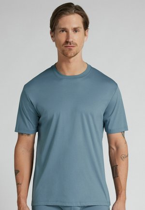 Mann mit kurzen braunen Haaren, der ein schlichtes hellblaues Kurzarm-T-Shirt trägt, steht vor einem neutral grauen Hintergrund.