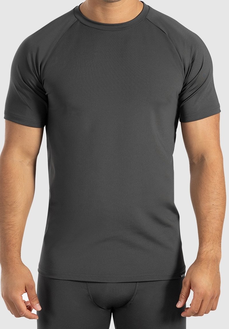 Homme portant un t-shirt de sport ajusté, à manches courtes, gris foncé et un short assorti, debout les bras détendus le long du corps devant un fond clair.