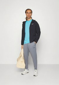 Nike-Sportoutfit mit einer schwarzen Jacke mit Reißverschluss, einem türkisfarbenen Shirt, grauen Hosen, weißen Sneakers mit grünen Akzenten und einer beigen Reisetasche.