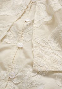 Tissu de couleur crème avec broderie florale, présentant des boutons blancs alignés verticalement le long de la couture centrale.